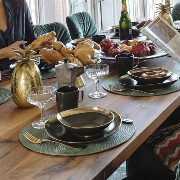 Der Frühstückstisch ist mit schwarzem und goldenem Geschirr, Gebäck, Obst und Getränken gedeckt; die Leute sitzen darum herum.