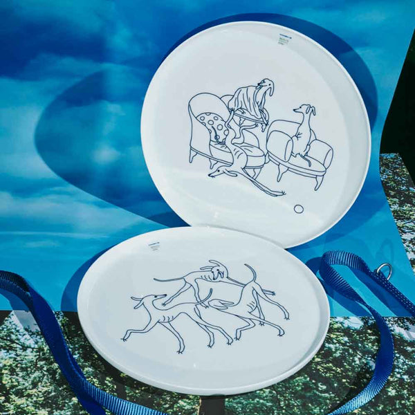 Zwei weiße Teller mit blauen Strichzeichnungen von Hunden, die auf Möbeln liegen und miteinander spielen, vor blauem Hintergrund.