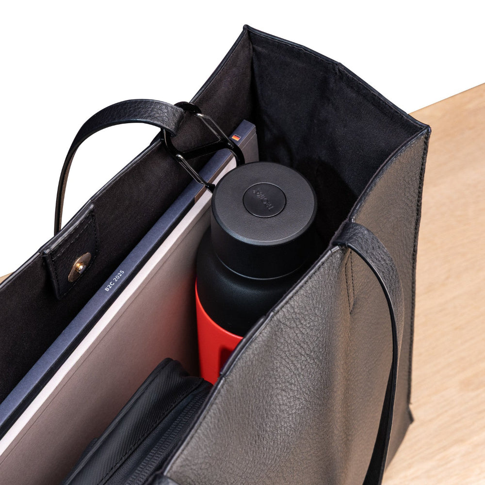Eine schwarze Tragetasche mit einer Wasserflasche, einem geschlossenen Laptop, einem Notizbuch und einer Tasche mit Reißverschluss, die auf einer hellen Holzoberfläche ruht.
