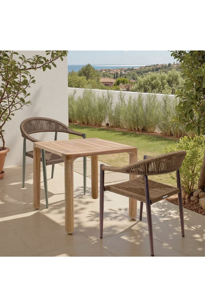 Zwei geflochtene Stühle und ein Holztisch stehen auf einer gefliesten Terrasse, im Hintergrund sind Pflanzen und ein Garten zu sehen.