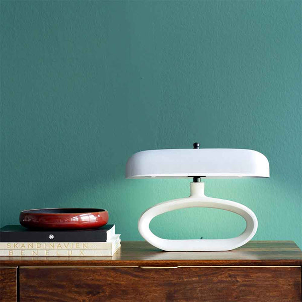 Eine moderne weiße Tischlampe und eine rote Schale stehen auf einem Bücherstapel auf einer hölzernen Oberfläche vor einer blaugrünen Wand.