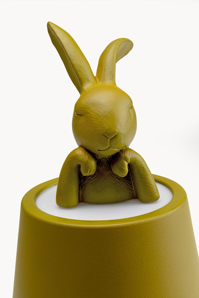 Eine gelblich-grüne stilisierte Kaninchenfigur mit geschlossenen Augen und erhobenen Pfoten sitzt auf einem passenden zylindrischen Sockel vor einem weißen Hintergrund.