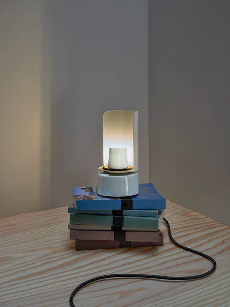 Eine moderne Lampe auf gestapelten Büchern mit einer schwarzen Schnur, platziert auf einem hellen Holztisch vor einer grauen Wand.