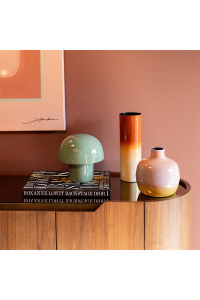 Auf einem hölzernen Sideboard stehen zwei Vasen, eine kleine grüne Lampe und ein Stapel Bücher unter einem gerahmten abstrakten Gemälde an einer rosa Wand.