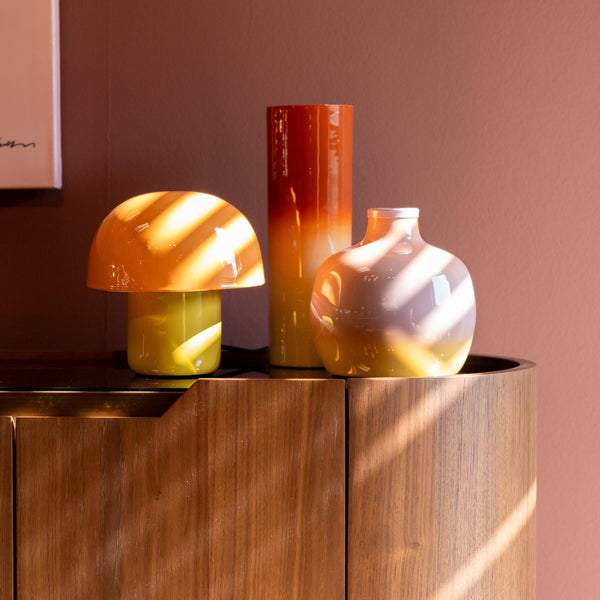 Drei orangefarbene und gelbe Dekorationsobjekte auf einem Holzschrank im Sonnenlicht vor einer rosa Wand.