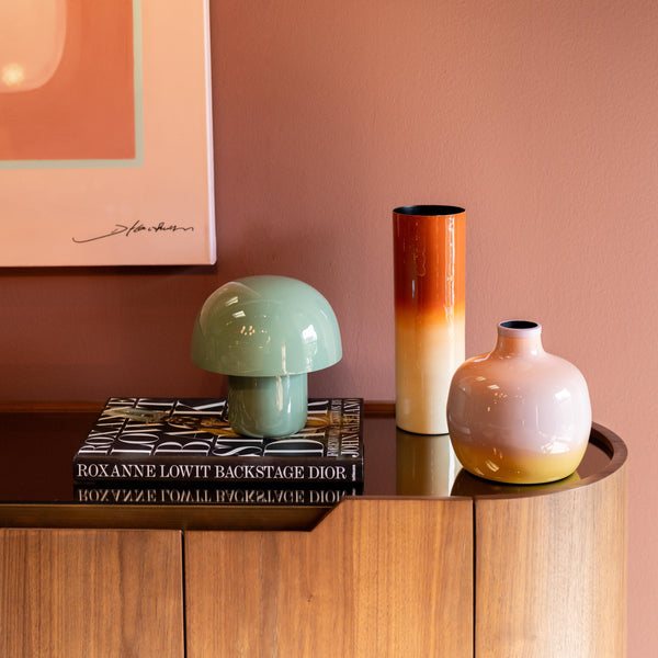 Ein hölzernes Sideboard mit Vasen, einer grünen Lampe, Kunstbüchern und rosafarbener Wandmalerei darüber.