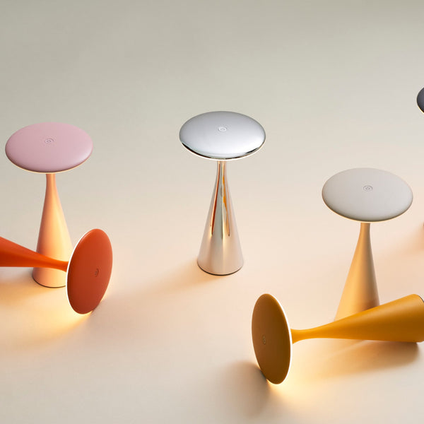 Eine Gruppe moderner, kegelförmiger Tischlampen in Silber, Rosa, Orange und Gelb, von denen einige aufrecht stehen und andere auf der Seite liegen, vor einem neutralen Hintergrund.