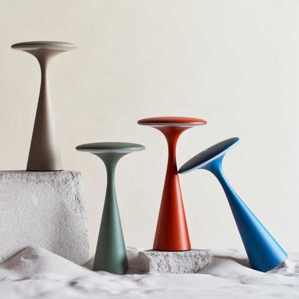 Vier moderne, pilzförmige Lampen in Beige, Grün, Rot und Blau sind auf strukturierten weißen Blöcken vor einem neutralen Hintergrund angeordnet.