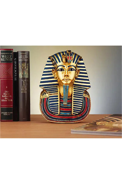 Ein Modell der goldenen Maske von König Tutanchamun steht auf einem Holzregal zwischen Büchern, darunter auch Bände zur Kunstgeschichte, wobei ein weiteres Buch teilweise im Vordergrund zu sehen ist.