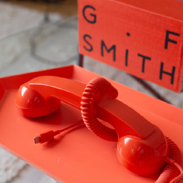 Ein roter Telefonhörer im Retrostil mit einem USB-C-Kabel liegt auf einem passenden roten Tablett neben einem roten Schild mit der Aufschrift G F SMITH.