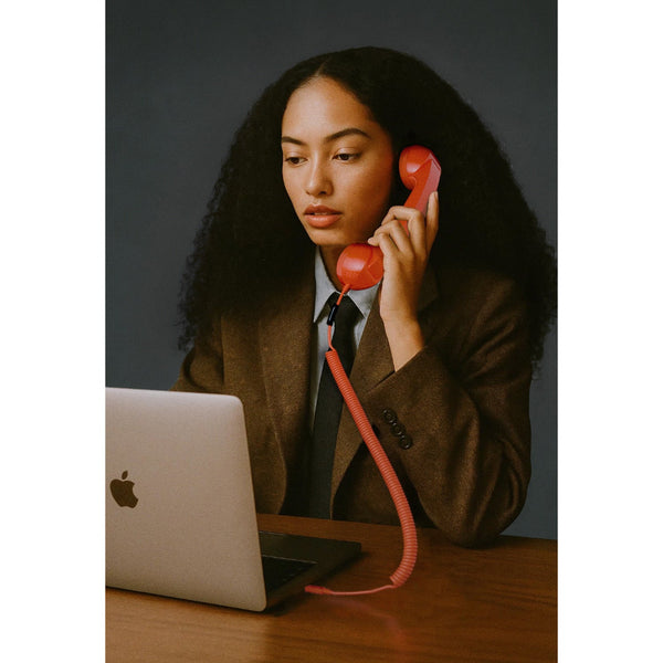 Eine Frau in einem braunen Blazer sitzt an einem Schreibtisch mit einem Laptop und hält ein rotes schnurgebundenes Telefon an ihr Ohr.