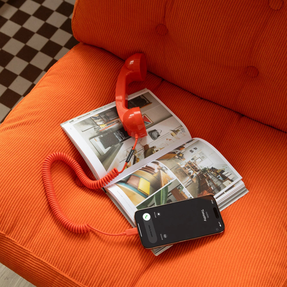 Auf einer orangefarbenen Couch liegen ein roter kabelgebundener Hörer und ein offenes Magazin; ein Smartphone, das einen eingehenden Anruf anzeigt, ist über ein Spiralkabel mit dem Hörer verbunden.