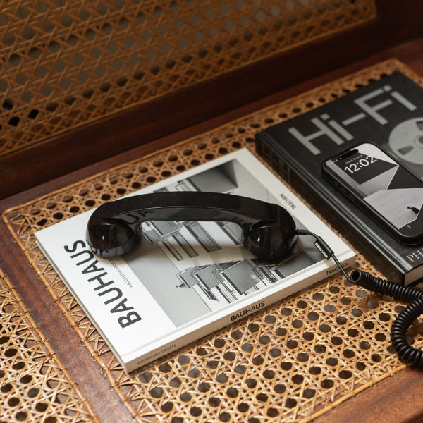 Ein schwarzer Vintage-Telefonhörer ruht auf einem Bauhaus-Buch neben einem Hi-Fi-Magazin und einem Smartphone, das die Uhrzeit 12:02 anzeigt, alles auf einer Oberfläche aus geflochtenem Schilfrohr.