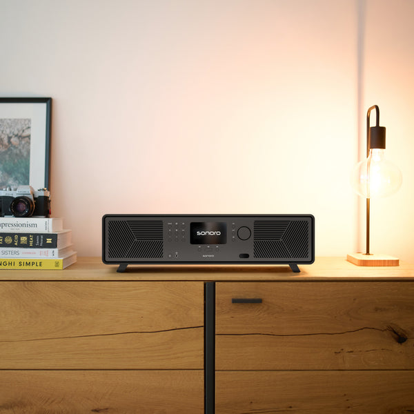 Schwarzes Sonoro-Audiosystem auf einem Holzschrank mit Büchern, gerahmtem Foto und Glühbirnenlampe.