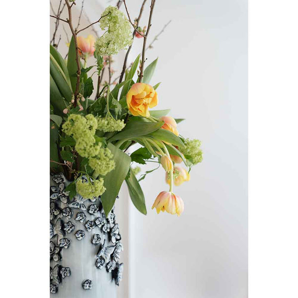 Gelbe Tulpen und grüne Blumen in einer strukturierten weißen Vase vor einem schlichten, hellen Hintergrund.