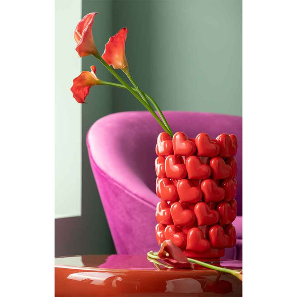 Rote herzförmige Vase mit orangefarbenen Calla-Lilien auf einem Tisch, lila Stuhl und grüne Wand im Hintergrund.