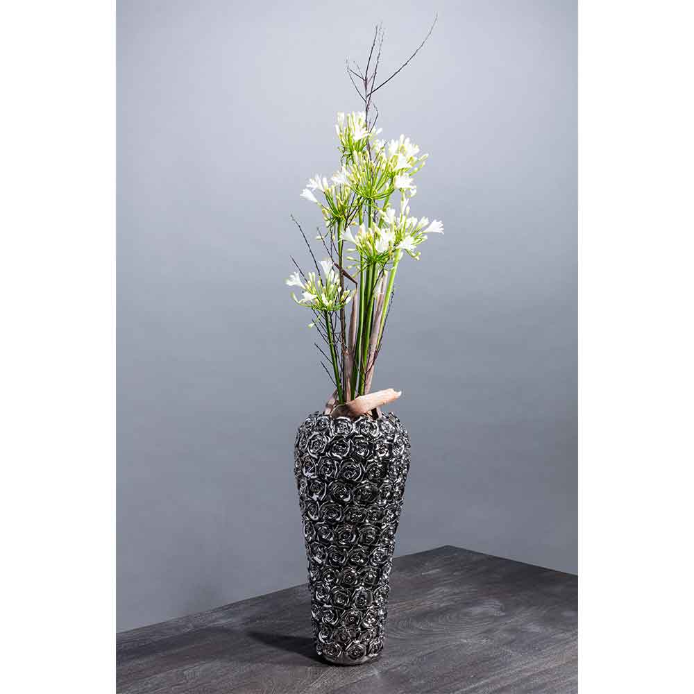 Hohe verzierte Vase mit weißen Blumen und dünnen Zweigen auf einem dunklen Tisch vor einem schlichten grauen Hintergrund.