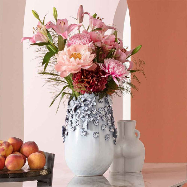 Eine Vase mit rosa Blumen auf einem Marmortisch, daneben Pfirsiche und ein weißer Zierkrug.