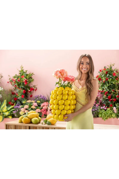 Eine Frau in einem gelben Outfit steht drinnen neben einem Tisch mit Zitronen und hält eine große dekorative Vase  aus Zitronen und rosa Blumen in der Hand. Im Hintergrund sind bunte Blumen zu sehen.