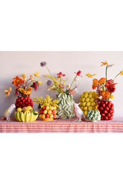 Ein Tisch mit Vogelfiguren aus Keramik, bunten Blumen und Obstarrangements, darunter Zitronen, Erdbeeren und Orangen, auf einer rosa gestreiften Tischdecke vor einer schlichten Wand.