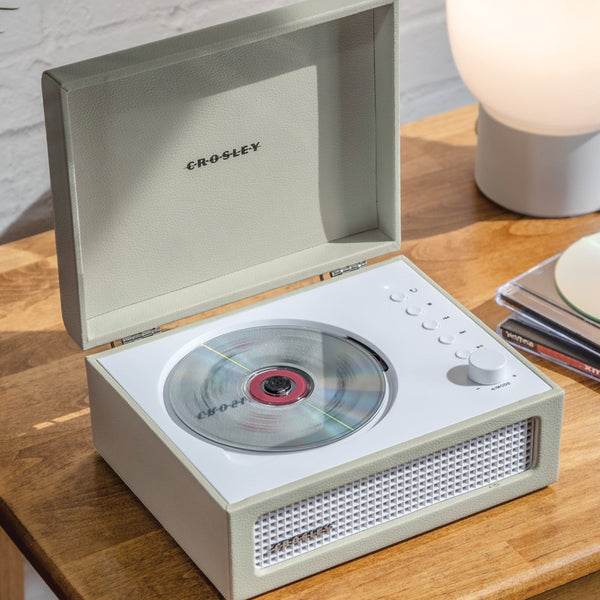 Ein Crosley-CD-Player mit offenem Deckel steht auf einem Holztisch und spielt eine CD ab. In der Nähe befinden sich gestapelte CDs und eine runde weiße Lampe.