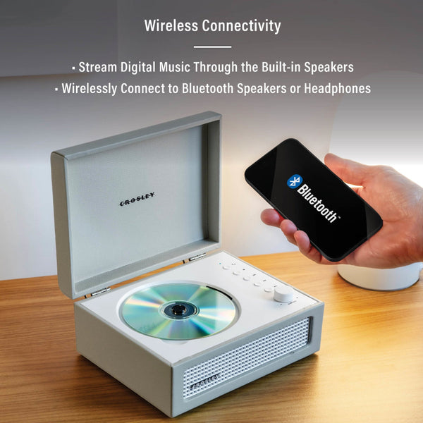 Eine Person hält ein Smartphone mit einem Bluetooth-Symbol in der Nähe eines Crosley-CD-Players mit eingebauten Lautsprechern auf einem Holztisch, um die drahtlosen Verbindungsfunktionen hervorzuheben.