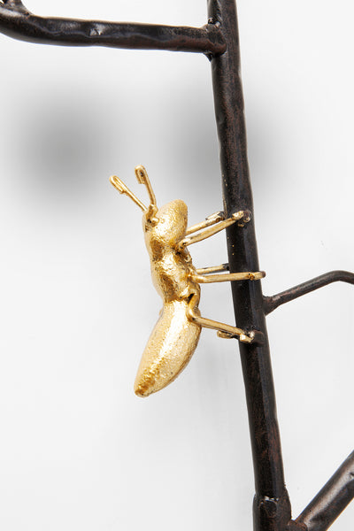 Eine goldfarbene Ameisenskulptur, die auf einem dunklen Metallzweig vor einem weißen Hintergrund klettert.