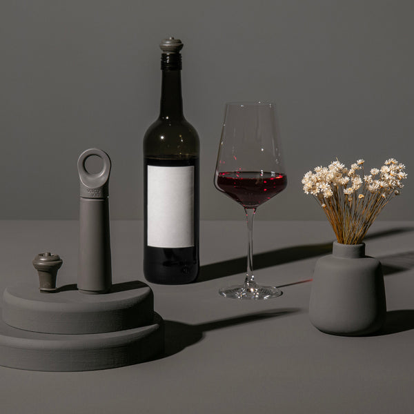 Eine Weinflasche, ein Glas Rotwein und eine graue Vase mit weißen Blumen auf einer grauen Fläche mit gedämpfter Beleuchtung.