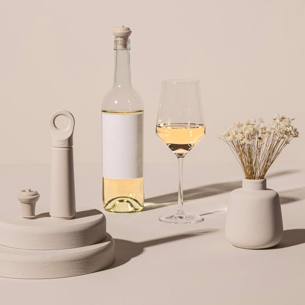 Eine Flasche Weißwein, ein gefülltes Weinglas, Keramikdekor und eine kleine Vase mit Trockenblumen auf einer beigen Fläche.