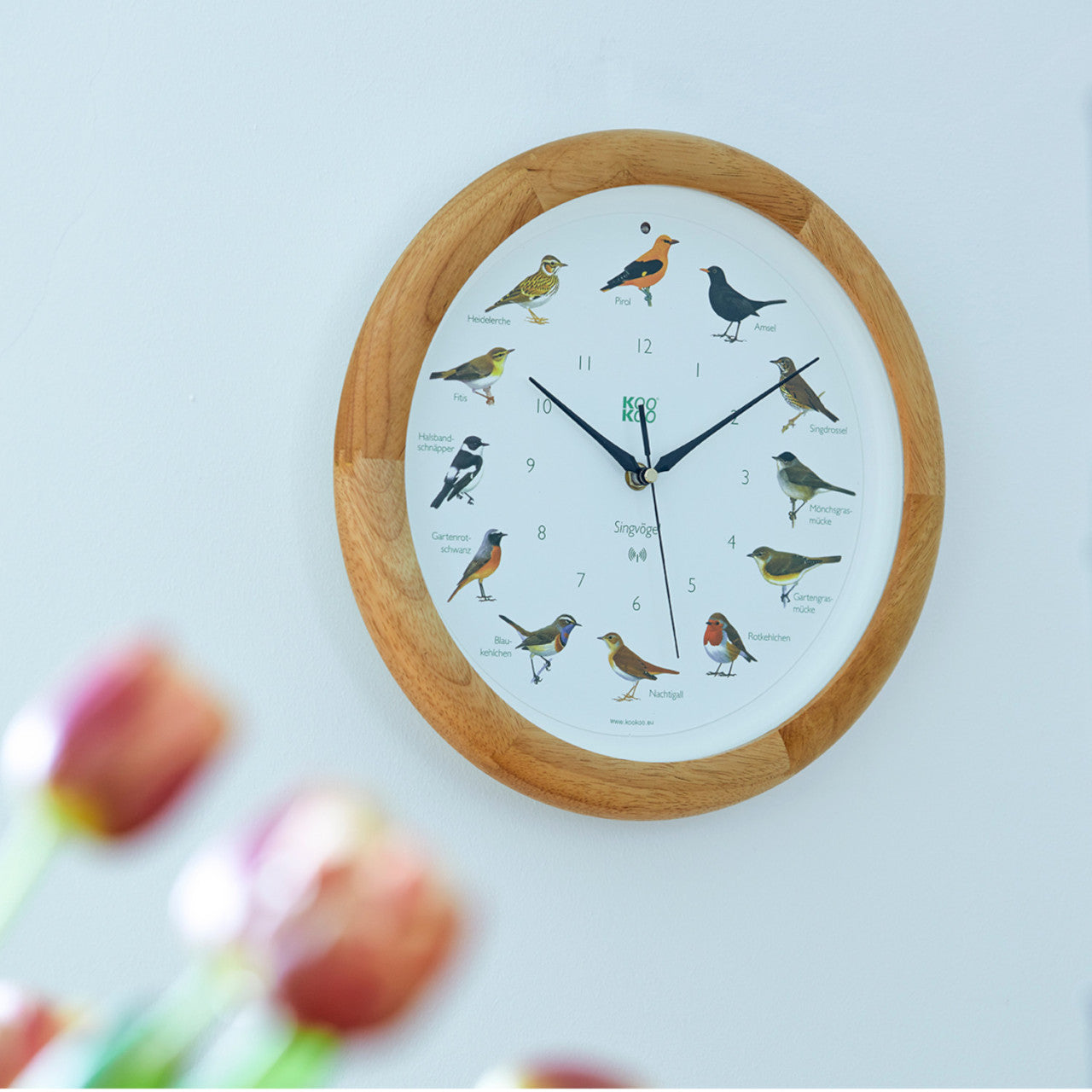 Die KooKoo Wanduhr Singvögel – Holz ist eine runde Wanduhr mit Vogelillustrationen zu jeder vollen Stunde und verschwommenen Blumen im Vordergrund.