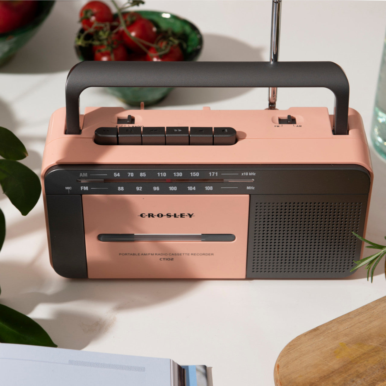 Ein Crosley-Kassettenspieler in Rosa/Grau steht auf einem Tisch neben Pflanzen und Tomaten.