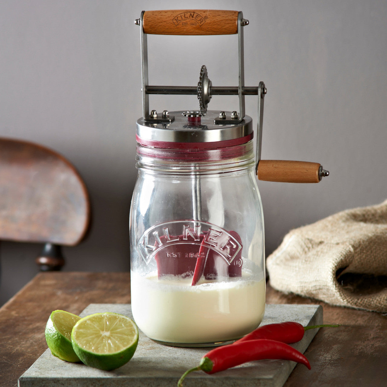Das Butter Fass 1 Liter, ein Butterfass aus Glas mit Holzgriff, steht neben Limettenhälften und roten Chilischoten auf einem Tisch.