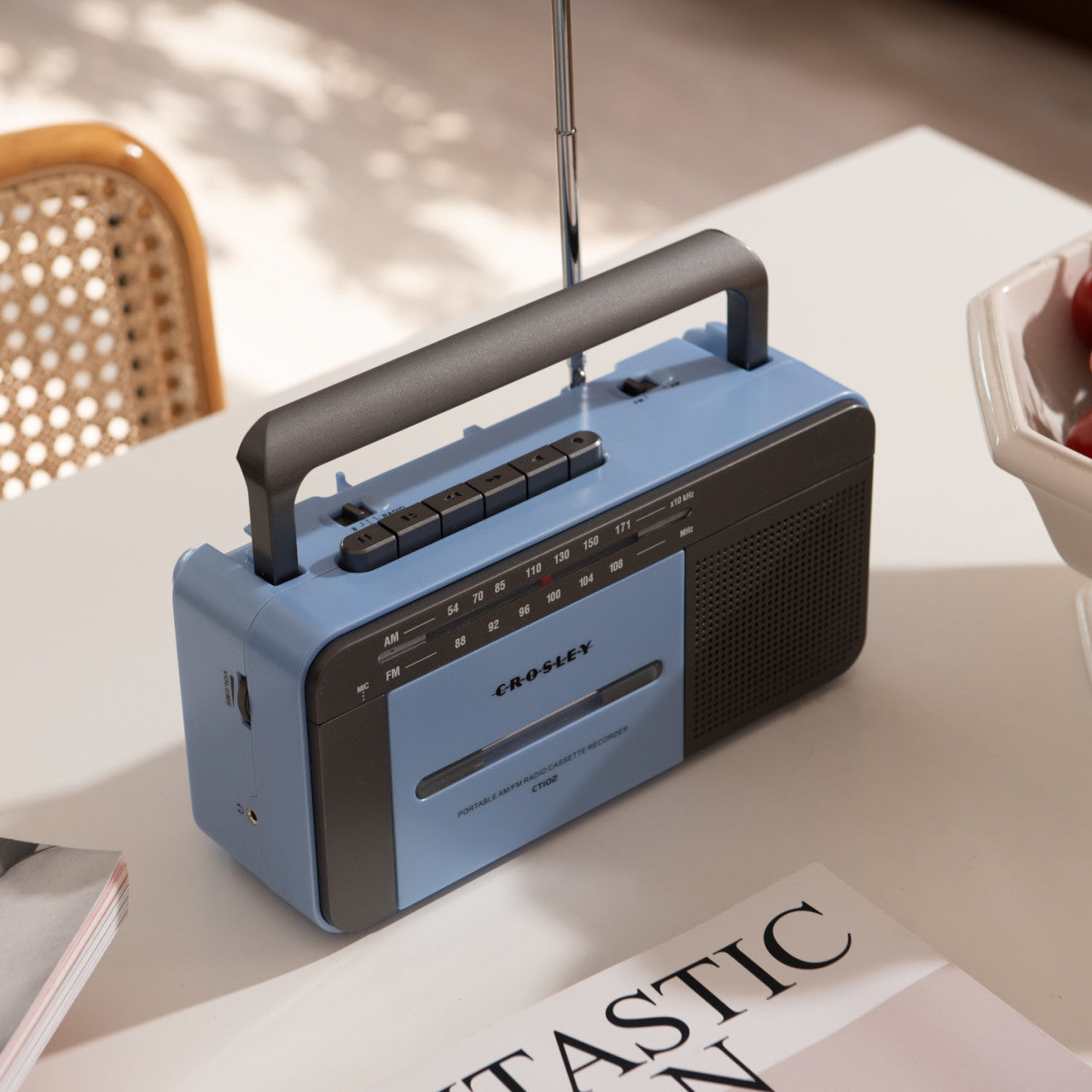 Ein blau-grauer Crosley-Kassettenspieler steht auf einem weißen Tisch neben einer Zeitschrift und einer Schale Äpfel.