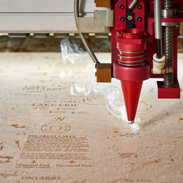 Ein Laserschneider ätzt detaillierten Kartentext in eine flache Holzoberfläche, wobei aus der Gravurstelle Rauch aufsteigt.