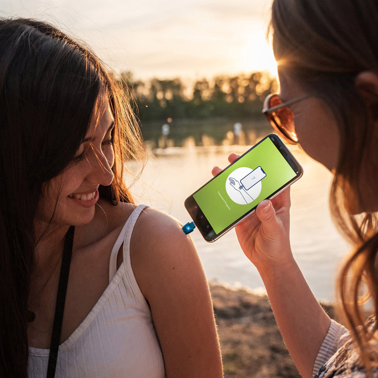 Zwei Frauen lächeln im Freien an einem See bei Sonnenuntergang; die eine behandelt mit ihrem Smartphone und dem Insektenstichheiler heat it classic Lightning den Arm der anderen.