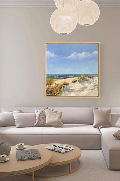 Gemälde eines Sandstrandes mit grasbewachsenen Dünen im Vordergrund, Wellen im Meer und einem blauen Himmel mit ein paar Wolken, gerahmt in hellem Holz - in einem Wohnzimmer über einem Sofa hängend. 