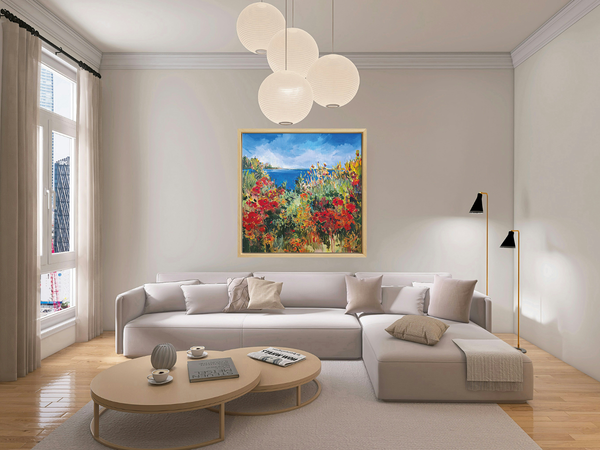 Gerahmtes Gemälde einer lebendigen Küstenlandschaft mit roten Blumen, grünem Laub und einem blauen Himmel über einem Gewässer - an einer Wand über einem Sofa hängend. 