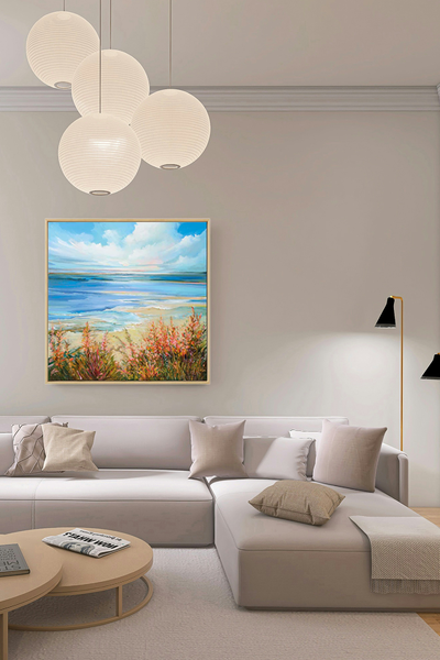 Ein gerahmtes Bild zeigt einen Sandstrand mit orangefarbenen Blumen im Vordergrund, ruhiges blaues Wasser in der Mitte und einen hellen Himmel mit Wolken darüber - an einer Wohnzimmerwand hängend vor einem modernen Sofa und einem Couchtisch.