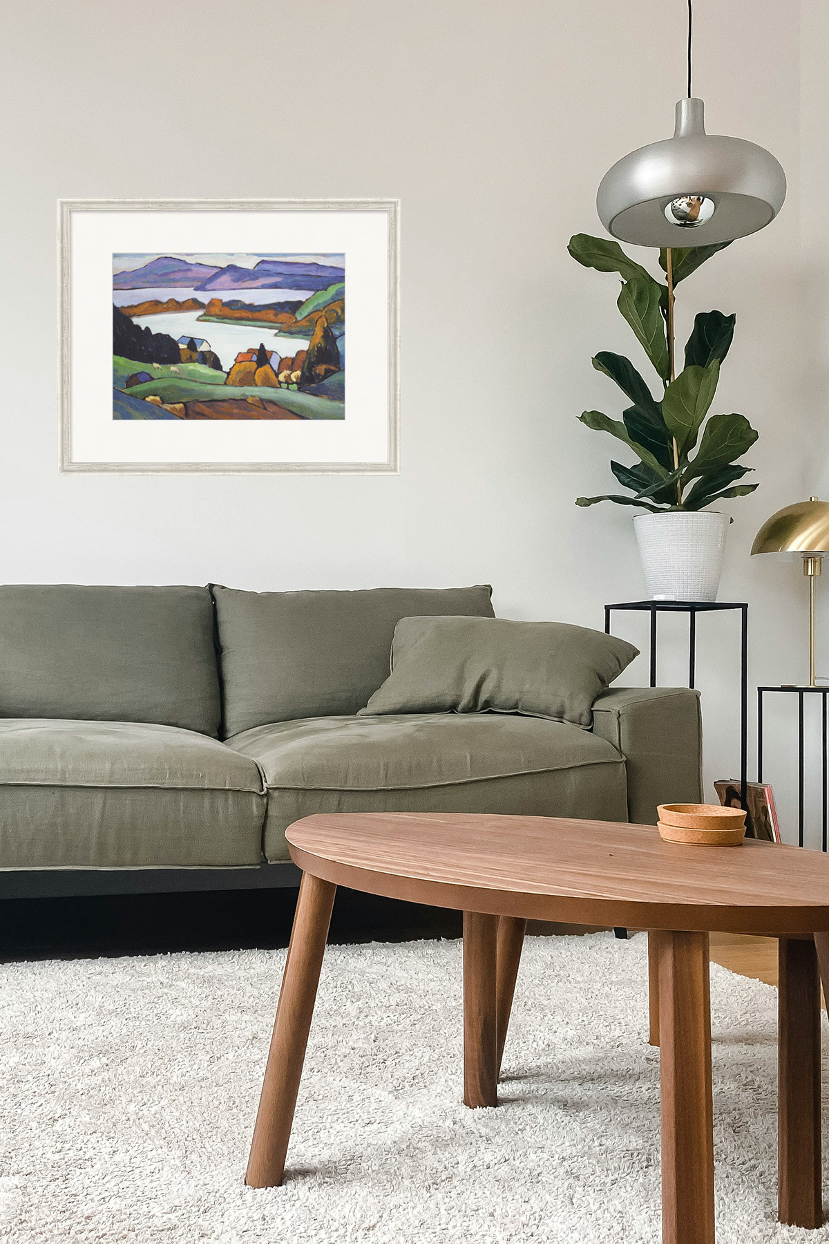 Farbenfrohes Gemälde eines Dorfes am See mit Bergen im Hintergrund, weiß gerahmt -  in einem Wohnzimmer mit grüner Couch an der Wand hängend. 