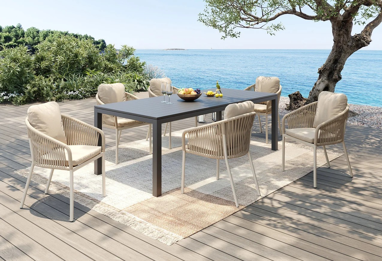 Erleben Sie Entspannung am Meer mit dem Stuhl Cyprus Beige Set – sechs gepolsterte Stühle mit Aluminiumgestell und Polypropylen-Geflecht um einen rechteckigen Tisch auf einer Holzterrasse mit Blick aufs Meer. Perfekt für Mahlzeiten im Freien.