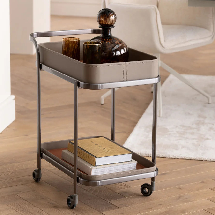 Ein Barwagen mit Gläsern auf der Oberseite und Büchern auf dem unteren Regal, auf einem Holzboden neben einem Teppich.