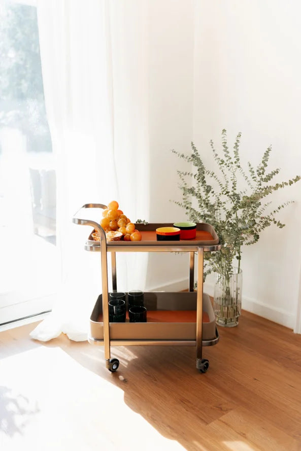 Ein Barwagen mit Orangen, Gläsern und einer Vase mit Grünzeug steht an einem sonnenbeschienenen Fenster auf einem Holzboden.