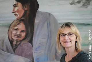 Eine Frau mit Brille lächelt neben „Anja Struck: 4 Bilder ‚Glücksmomente‘ (2024) im Set, gerahmt“, auf dem ein Gemälde einer Frau und eines Kindes zu sehen ist, die in eine Decke gehüllt sind.