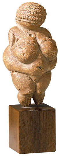 Steinerne Venusfigur mit übertriebenen Gesichtszügen, auf einem Holzsockel stehend, von vorne betrachtet.