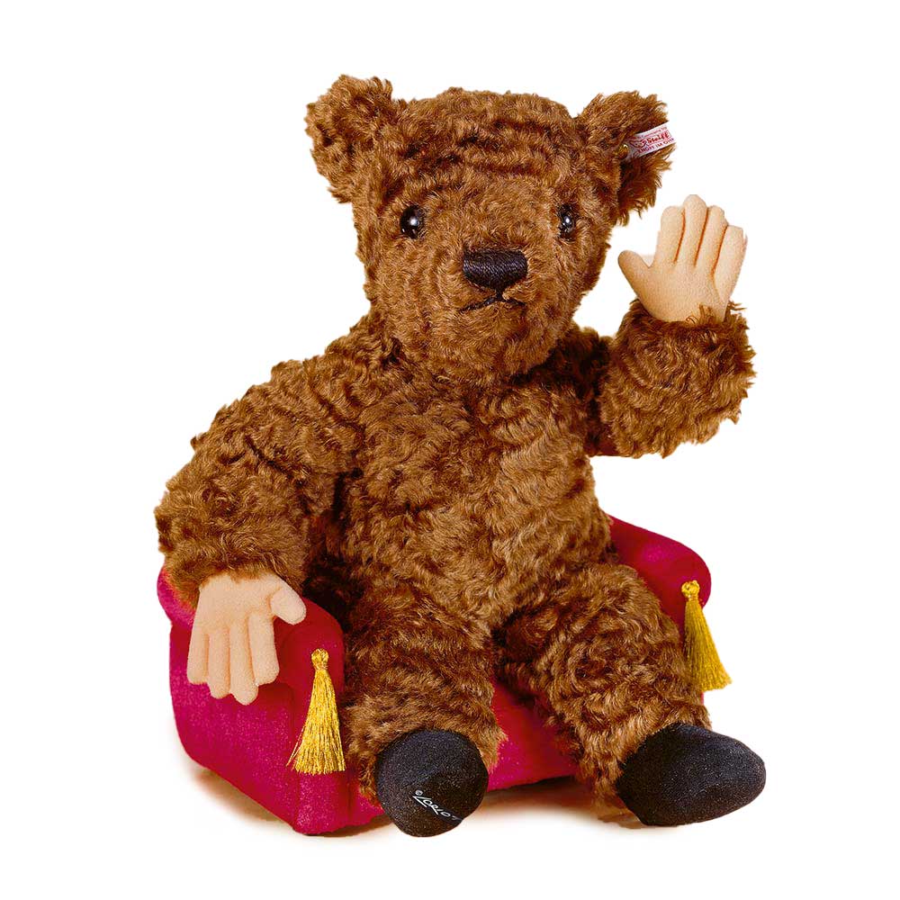 Ein brauner Teddybär winkt, während er auf einem kleinen roten Sessel mit gelben Quasten sitzt.