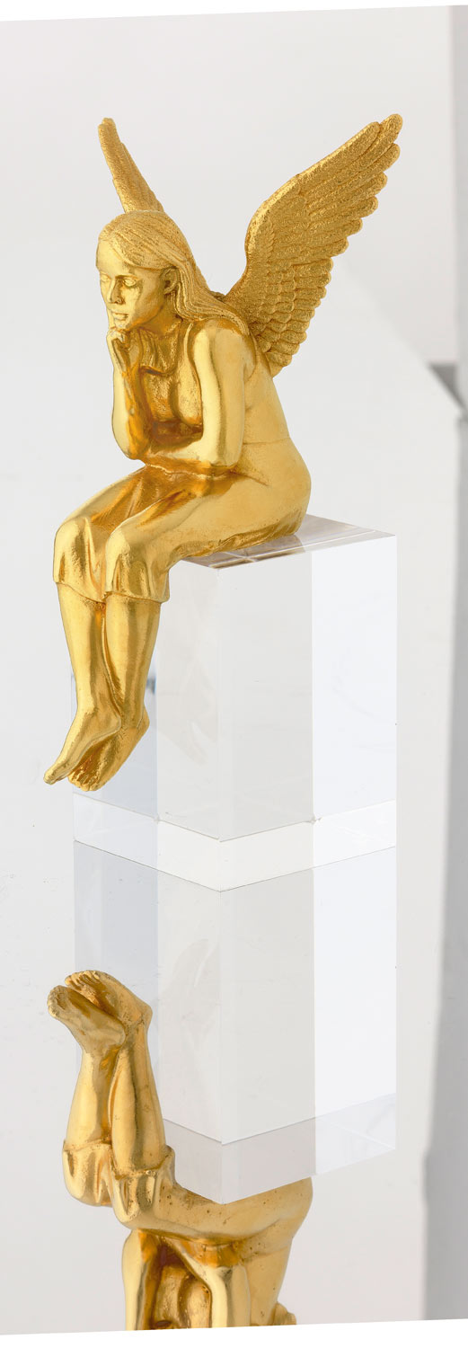 Eine goldene Engelsfigur sitzt nachdenklich auf einem durchsichtigen Block, darunter ist ihr Spiegelbild sichtbar.