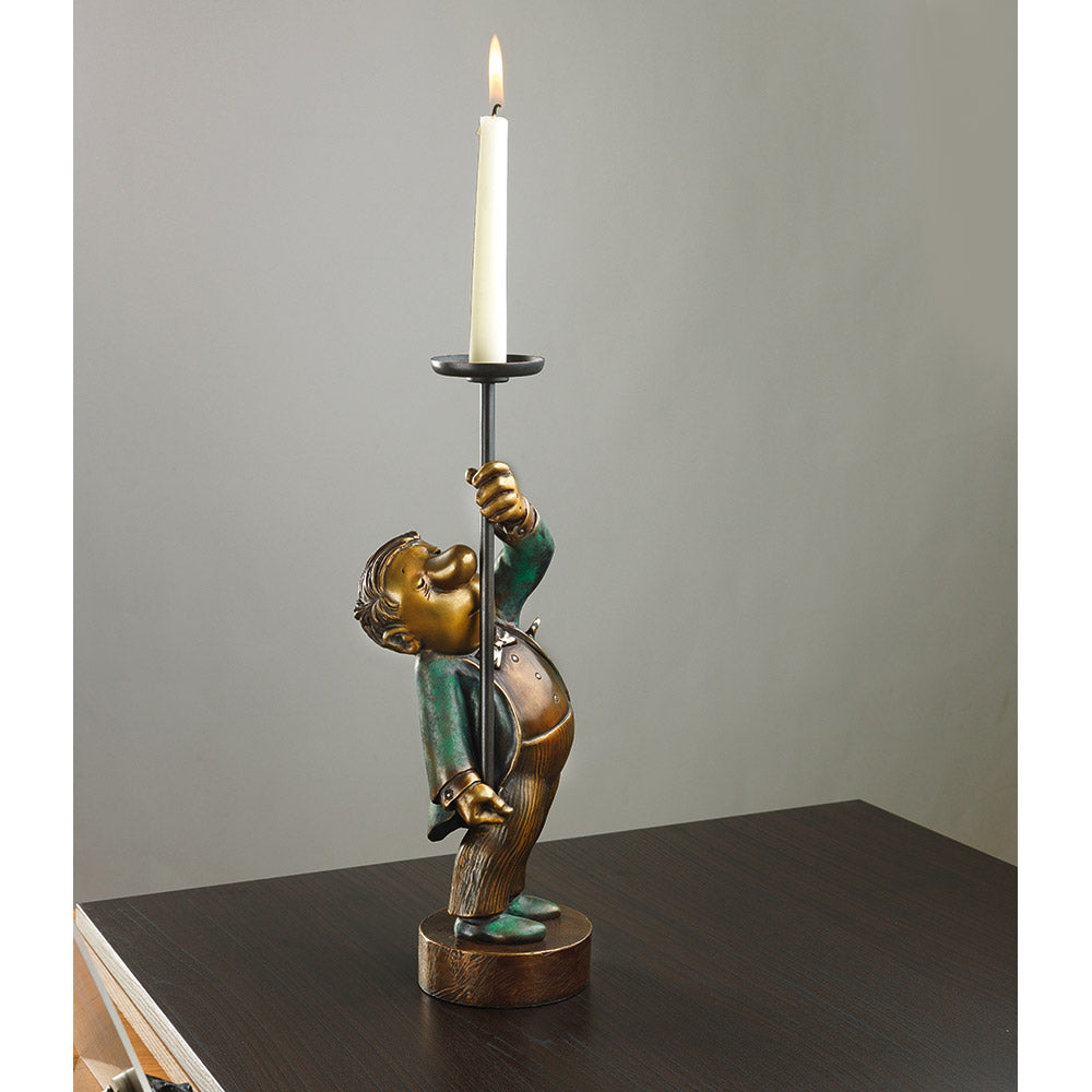 Eine skurrile Figur, die eine große Kerze hält und auf einem dunklen Holztisch vor einem schlichten Hintergrund steht.