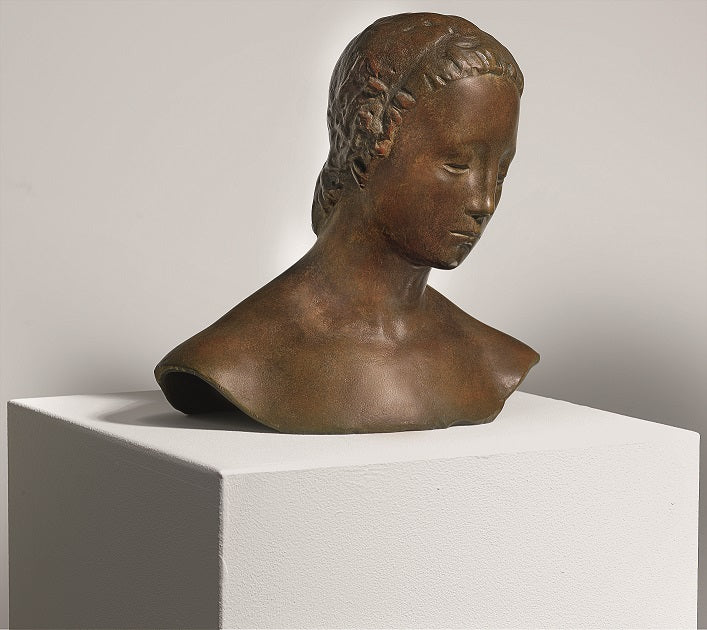 Bronzebüste einer jungen Frau mit geflochtenem Haar auf einem weißen Sockel vor schlichtem Hintergrund.