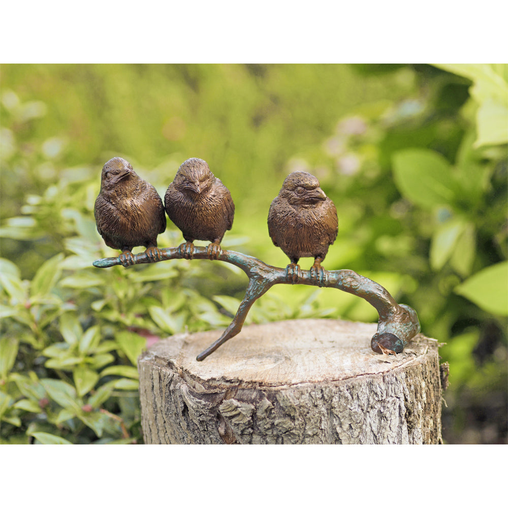 Drei Vogelskulpturen sitzen auf einem Ast auf einem Baumstumpf, im Hintergrund ist Grün.