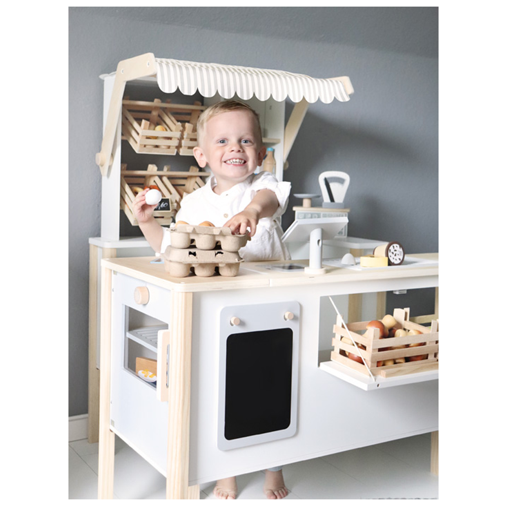 Ein lächelndes Kind spielt am hölzernen Marktstand „Kaufladen und Kinderküche fresh“, hält Eier in der Hand und ist von Spiellebensmitteln umgeben.